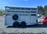 Side view of a white livestock trailer with a spare tire mounted on the door, tandem axles, and a front hitch on a gravel lot with trees in the background.