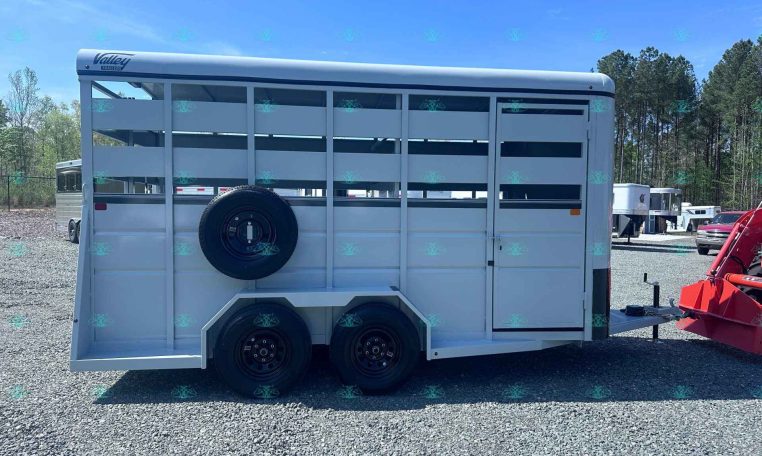 Side view of a white livestock trailer with a spare tire mounted on the door, tandem axles, and a front hitch on a gravel lot with trees in the background.