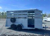 Two-axle aluminum horse trailer with open side doors, spare tire mounted on the left, parked on a gravel lot under a blue sky.