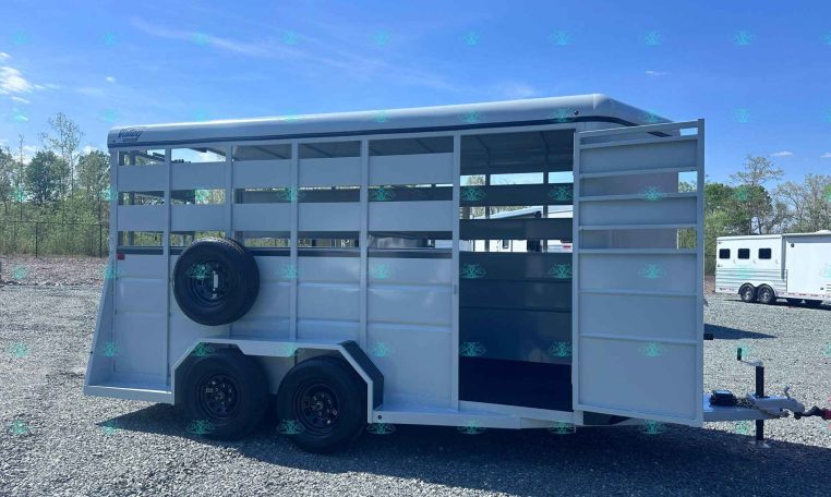 Two-axle aluminum horse trailer with open side doors, spare tire mounted on the left, parked on a gravel lot under a blue sky.