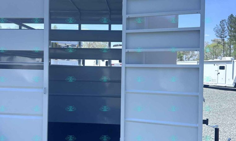 White trailer with back door open, showing interior shelves and a dark divider, parked on gravel with blue sky above.