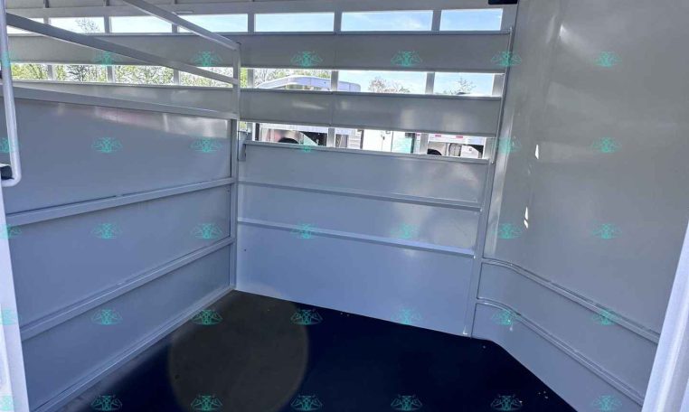 Interior of a light gray horse trailer with partitioned stalls, metal walls, and a black rubber floor; ventilation slots along the sides.