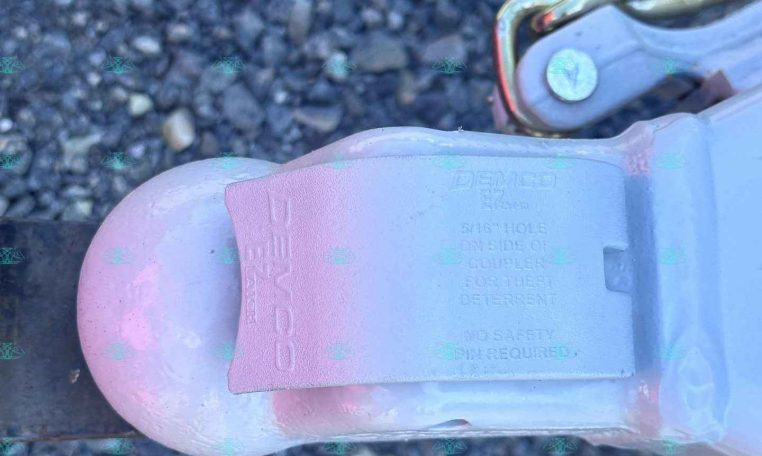 Close-up of a trailer hitch coupler with a safety pin and chain on gravel, showing Demco branding on the latch label.