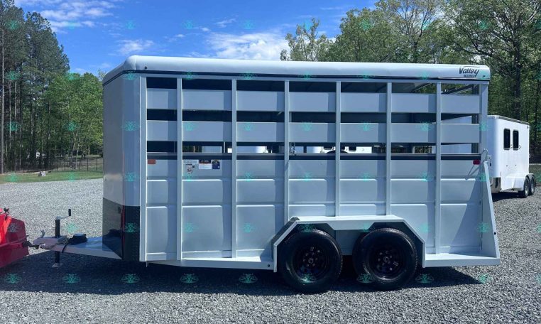 White tandem-axle livestock trailer on a gravel lot with trees in the background; CarolinaTrailerSales watermark at bottom.