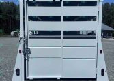 Rear view of a white horse trailer with split doors open, showing interior stalls on a gravel lot under a blue sky.