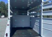 Open horse trailer interior with a center divider, empty stalls, and gray walls under a clear blue sky on a gravel lot.