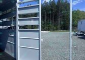 Silver aluminum livestock trailer with partitioned stalls and rear ramp open, on a gravel lot with trees in the background