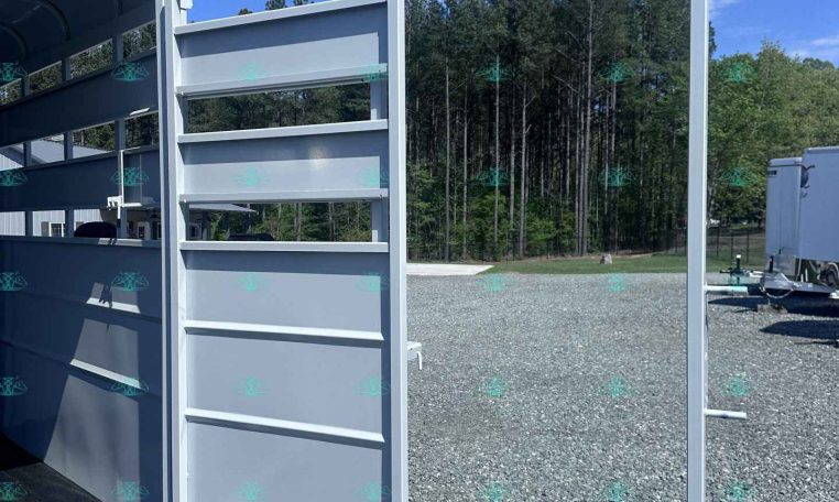 Silver aluminum livestock trailer with partitioned stalls and rear ramp open, on a gravel lot with trees in the background