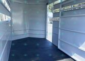 Interior of an empty white horse trailer with a black rubber floor and open back ramp, daylight outside visible
