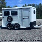 White horse trailer with dual axles and a spare tire mounted on the back, parked on a gravel lot with trees in the background.