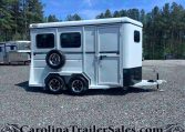 White horse trailer with dual axles and a spare tire mounted on the back, parked on a gravel lot with trees in the background.