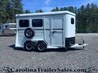 White horse trailer with dual axles and a spare tire mounted on the back, parked on a gravel lot with trees in the background.
