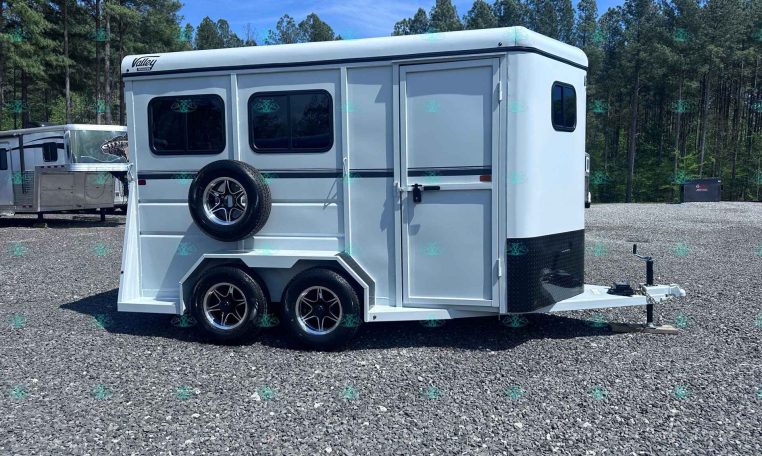 White horse trailer with dual axles and a spare tire mounted on the back, parked on a gravel lot with trees in the background.