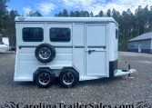 White enclosed horse trailer with a side-mounted spare tire, two windows, a side entry door, and a front hitch parked on a gravel lot with trees in the background.