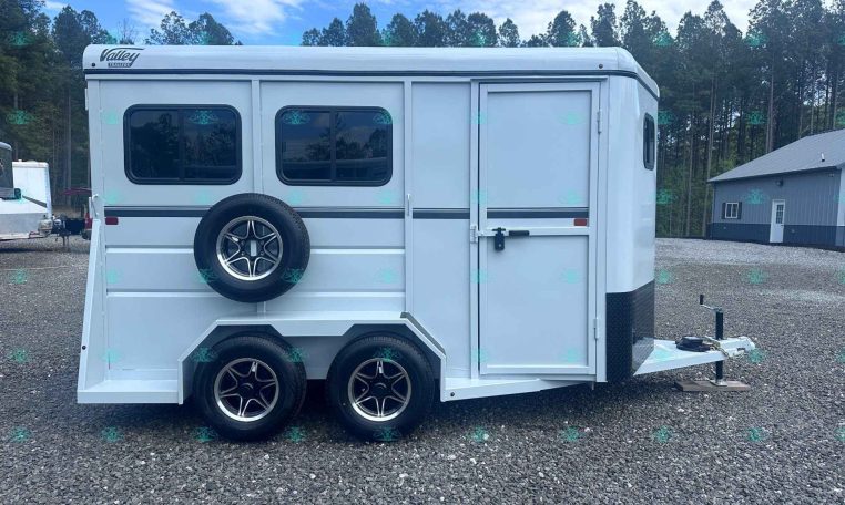 White enclosed horse trailer with a side-mounted spare tire, two windows, a side entry door, and a front hitch parked on a gravel lot with trees in the background.