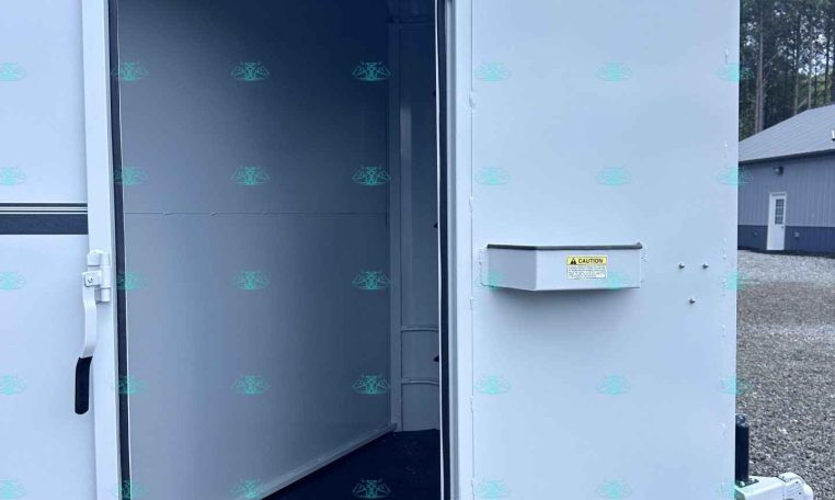 Open white enclosed cargo trailer with the rear door swung open, revealing a clean, empty interior with a dark floor against a gravel lot and blue sky.