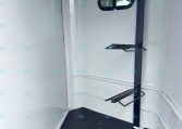 Interior of a trailer corner with a dark blue vertical support beam and two fold-down metal shelves attached, plus a small window and a non-slip black floor.