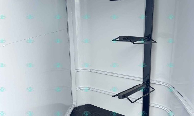 Interior of a trailer corner with a dark blue vertical support beam and two fold-down metal shelves attached, plus a small window and a non-slip black floor.