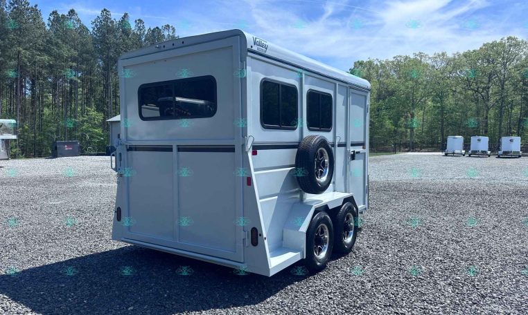 White horse trailer with two axles and spare tire mounted on the side, parked on a gravel lot with trees in the background.