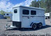 White enclosed horse trailer with two axles parked on a gravel lot, hitch and front guard visible, trees in the background.