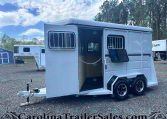 White, dual-axle horse trailer with side entry door open and barred windows, parked on gravel beside another trailer and a small blue shed. The CarolinaTrailerSales branding is visible at the bottom.