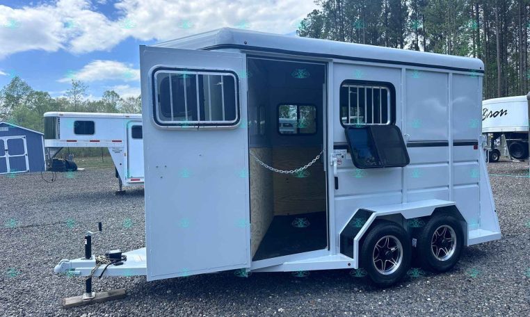 White, dual-axle horse trailer with side entry door open and barred windows, parked on gravel beside another trailer and a small blue shed. The CarolinaTrailerSales branding is visible at the bottom.
