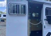 White horse trailer with open door, showing interior divider chain and barred window panels on a gravel lot.