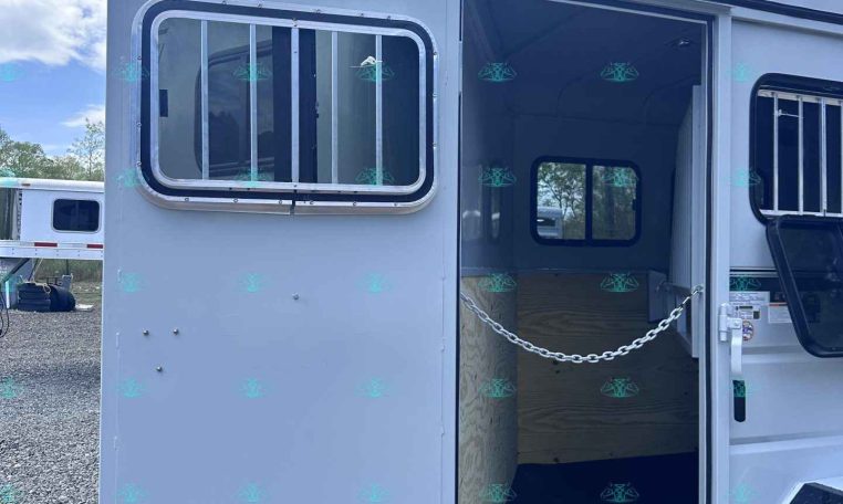 White horse trailer with open door, showing interior divider chain and barred window panels on a gravel lot.