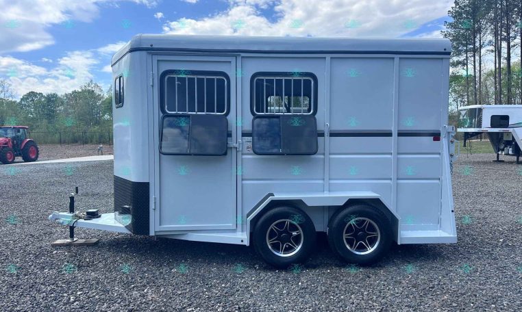 Side view of a light-gray, two-axle horse trailer with barred windows, parked on a gravel lot with trees in the background and a hitch at the front.