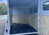 Empty white horse trailer interior with wooden side walls, divider, and open rear ramp on a gravel lot