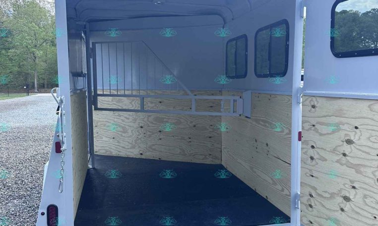 Empty white horse trailer interior with wooden side walls, divider, and open rear ramp on a gravel lot