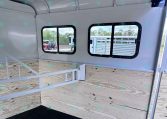 Interior of a light gray horse trailer with two side windows, wood-paneled walls, a white divider rail, and a black rubber floor. Outside equipment is visible through the windows.