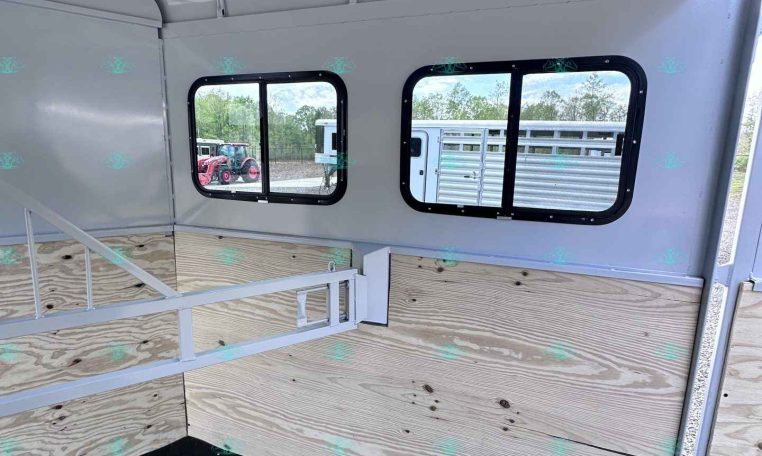 Interior of a light gray horse trailer with two side windows, wood-paneled walls, a white divider rail, and a black rubber floor. Outside equipment is visible through the windows.