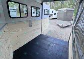 Interior of a light gray trailer with plywood side walls, a black rubber floor, and several windows along the left wall; rear ramp is open to a gravel lot with other trailers visible outside.