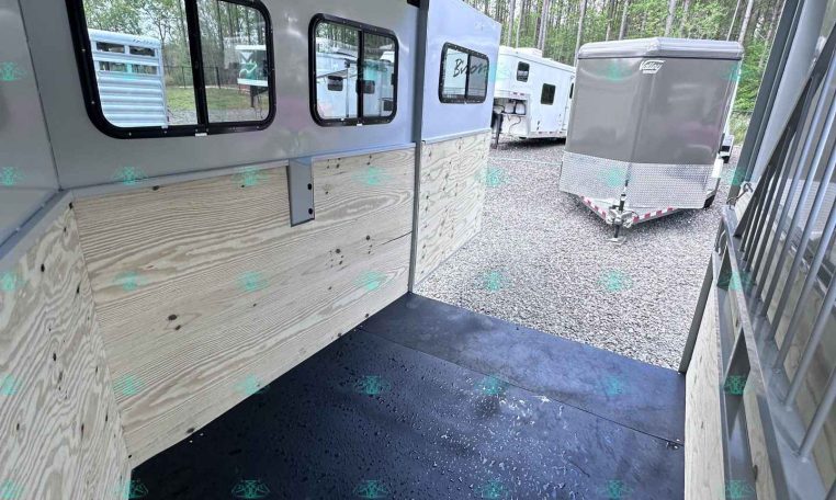 Interior of a light gray trailer with plywood side walls, a black rubber floor, and several windows along the left wall; rear ramp is open to a gravel lot with other trailers visible outside.