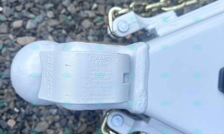 Close-up of a trailer coupler with safety chains on gravel, showing a Demco EZ-Latch and a 5/16 inch hole at the side of the coupler.