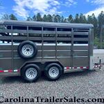 Gray livestock trailer with side vents and a mounted spare tire on the mid-side, parked on a gravel lot with trees in the background