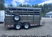 Gray livestock trailer with side vents and a mounted spare tire on the mid-side, parked on a gravel lot with trees in the background