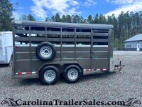 Gray livestock trailer with side vents and a mounted spare tire on the mid-side, parked on a gravel lot with trees in the background