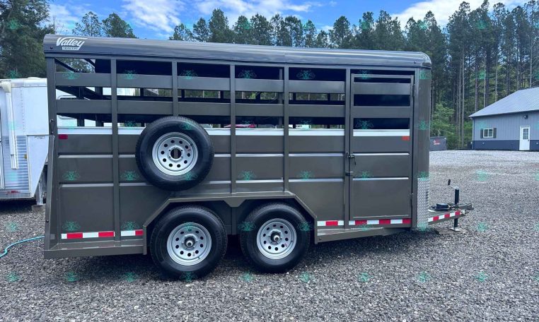 Gray livestock trailer with side vents and a mounted spare tire on the mid-side, parked on a gravel lot with trees in the background