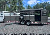 Gray dual-axle horse trailer with a side ramp open, spare tire mounted on the side, parked on gravel with trees in the background.