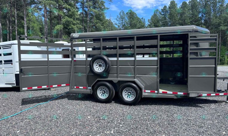 Gray dual-axle horse trailer with a side ramp open, spare tire mounted on the side, parked on gravel with trees in the background.