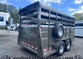 Gray metal livestock trailer with open slatted sides, two axles, and a spare tire mounted on the side; Value Trailers logo on top corner, parked on gravel ground.