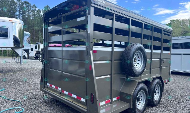 Gray metal livestock trailer with open slatted sides, two axles, and a spare tire mounted on the side; Value Trailers logo on top corner, parked on gravel ground.