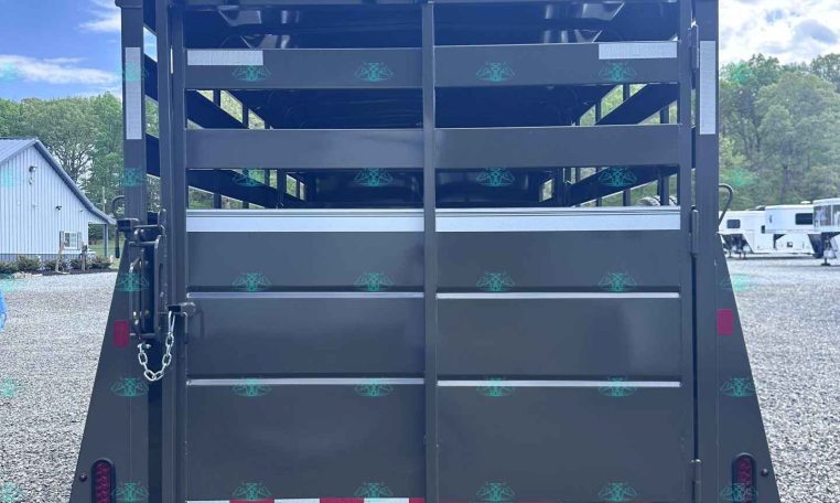 Rear view of a dark gray horse trailer with its rear doors open, revealing empty stalls and a wooden ramp on a gravel lot under a blue sky with clouds.