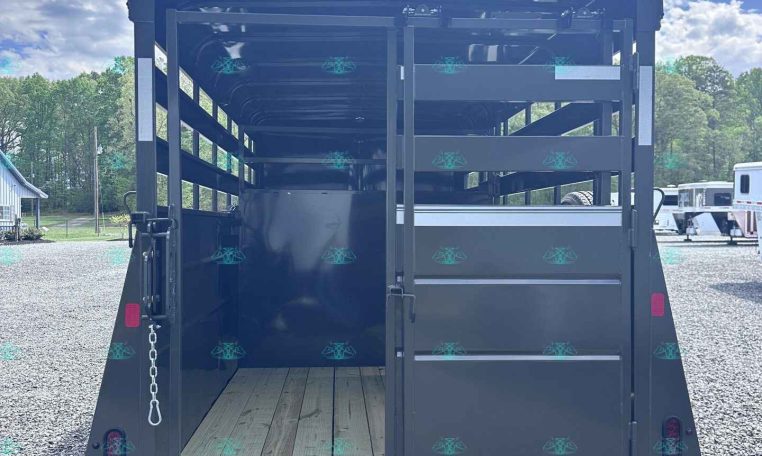 Back of a black horse trailer with interior stalls and a wooden floor, open rear doors, parked on gravel under a blue sky.