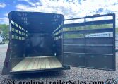 Open black livestock trailer with wooden floor and side rails, doors swung open on a gravel lot under a blue, cloudy sky.