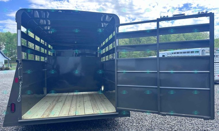 Open black livestock trailer with wooden floor and side rails, doors swung open on a gravel lot under a blue, cloudy sky.