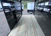 Interior of a black trailer with a wooden plank floor, view toward the back where another trailer is visible outside in a gravel lot.