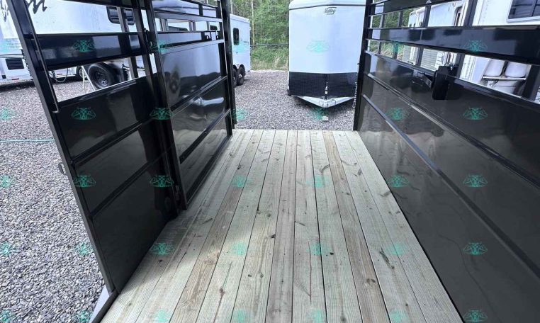Interior of a black trailer with a wooden plank floor, view toward the back where another trailer is visible outside in a gravel lot.